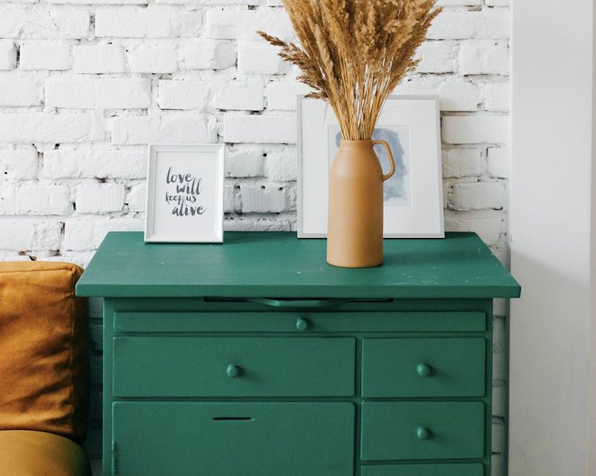 Green dresser with framed art and a vase of dried flowers against a white brick wall